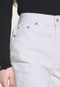 Valkoiset korkeavyötäröiset farkut, joissa korostetut saumat, metalliset ja kupariset niitit sekä sileä pinta. Edessä vetoketjusuljenta.