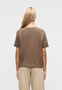 Ξανθός άνθρωπος, φαίνεται από πίσω, φοράει καφέ κοντομάνικο πουκάμισο και μπεζ παντελόνι, με φόντο ένα απλό ανοιχτό γκρι φόντο.