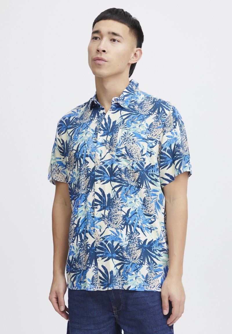 Camicia a maniche corte blu e crema con motivo tropicale di ananas e foglie, chiusura con bottoni sul davanti e dettaglio della tasca sul petto.