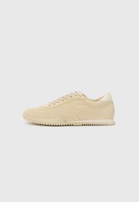 Beige suede sneaker with lace-up design, textured upper, contrasting rubber outsole, and a slight heel. Minimalist style with subtle branding.