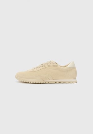 Sneaker beige in suede con design a lacci, parte superiore testurizzata, suola in gomma a contrasto e un leggero tacco. Stile minimalista con un branding discreto.