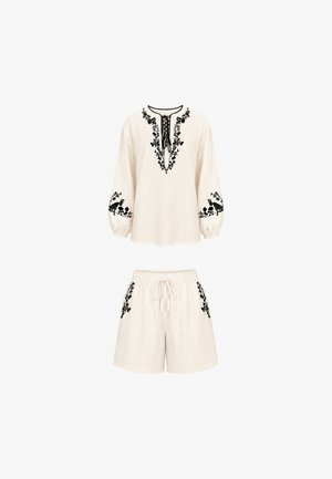 Completo crema con maglia a maniche lunghe e pantaloncini, ricamo floreale nero, scollo con laccio sulla maglia e girovita con coulisse sui pantaloncini.