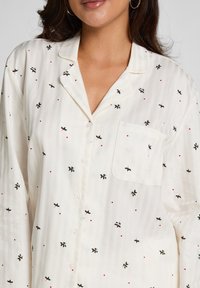 Weiß gestreiftes Schlafanzugoberteil mit grüner Stechpalmenblatt- und roter Beerenstickerei, ausgestattet mit einer Fronttasche und Knopfverschluss.