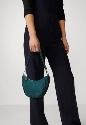 Persona con mangas largas negras y pantalones oscuros sosteniendo un pequeño bolso verde azulado con una correa corta a su lado.