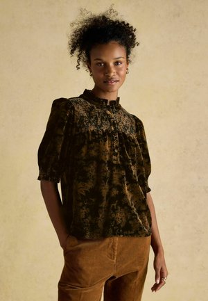 Dark green and gold velvet blouse with a ruffled neckline, short puffed sleeves, and floral pattern. Paired with brown corduroy pants.