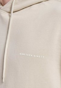 Beige Kapuzenpullover mit weißem Schriftzug "NINETEEN NINETY" auf der Vorderseite. Hergestellt aus weichem, strukturiertem Stoff.
