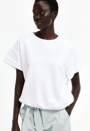 Frau trägt ein lockeres weißes Kurzarmshirt mit umgekrempelten Ärmeln und gestreifte hoch taillierte Hose, posiert mit einer Hand in der Tasche.