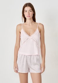 Młoda kobieta z prostymi czerwonymi włosami, nosząca jasnoróżową koszulkę na ramiączkach z koronkowym wykończeniem i różowe spodenki w paski, stojąca na tle białym.