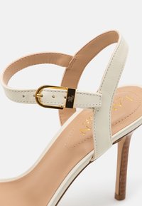 Białe skórzane sandały na wysokim obcasie z złotym paskiem z klamrą i brązową wyściółką, z wypukłym złotym napisem.