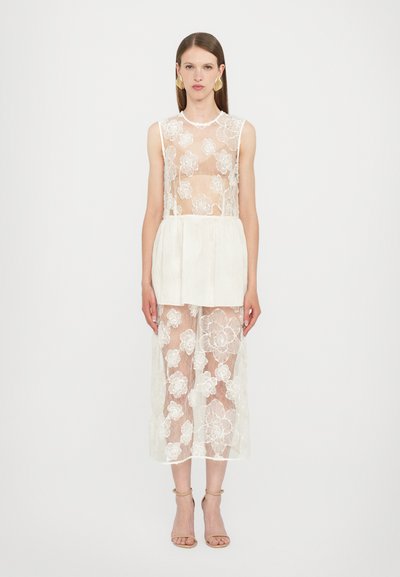 Witte kanten top met bloemenpatroon, laagjes over een crèmekleurige rok. Doorzichtige textuur, mouwloos ontwerp, gecombineerd met naakte strappy sandalen.