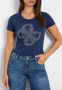 Tamsiai mėlynas medvilninis marškinėliai su dekoratyviniu sidabro spalvos krištolo grafikiniu dizainu. Trumpomis rankovėmis ir klasikinio apvalaus kaklo iškirpte. Dėvimi su mėlynomis džinsinėmis kelnėmis.