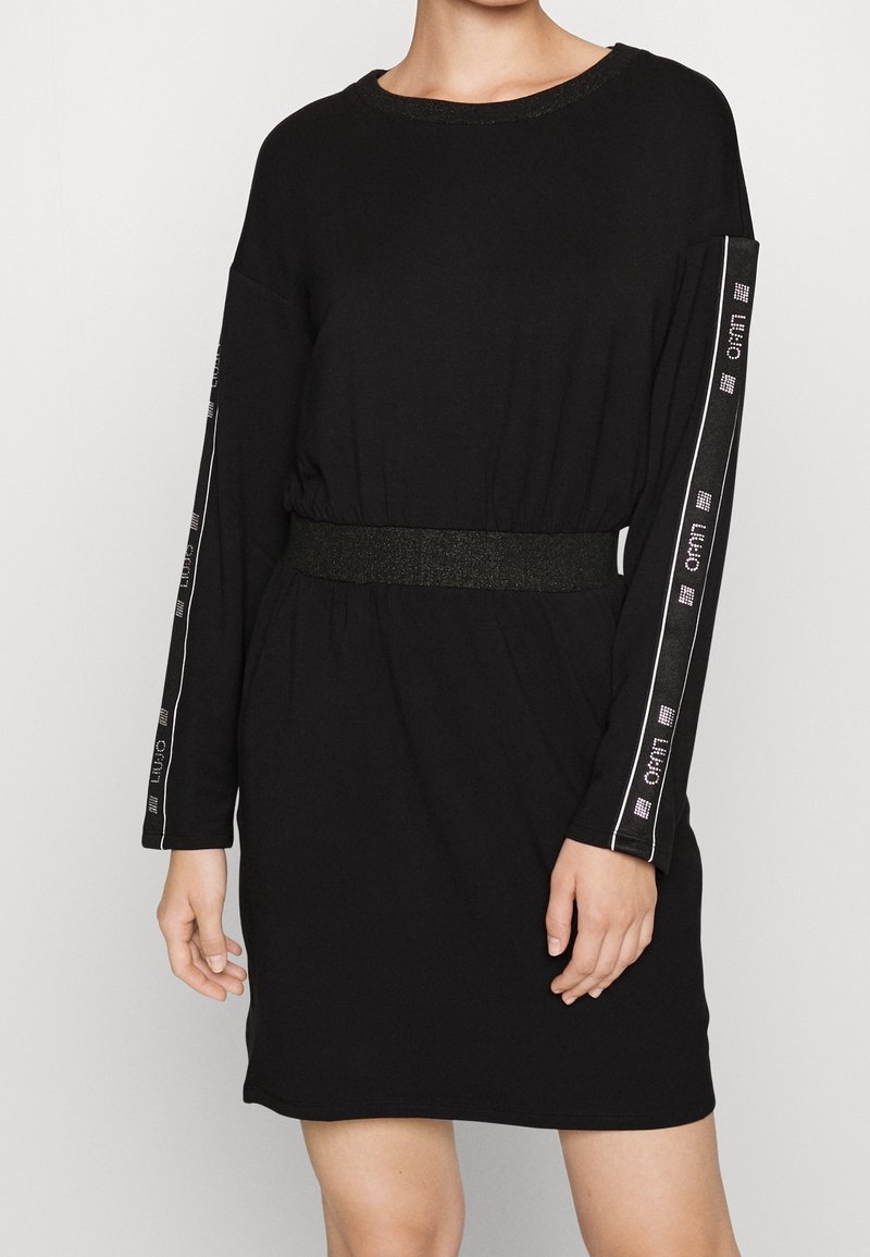 Femme portant une robe noire à manches longues avec une taille élastique et des bandes à logo courant le long des deux manches.