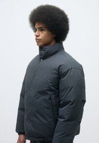 Puffer jacket in dark gray, featuring a high collar, button closure, and side pockets. Material appears durable and slightly textured.