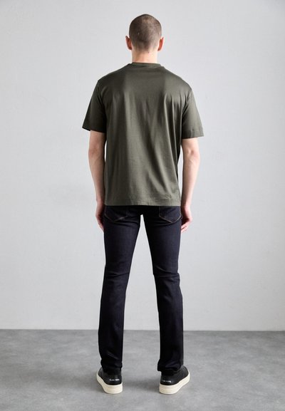 T-shirt en coton vert olive à manches courtes, associé à un jean en denim noir. Baskets noires avec une semelle blanche visibles aux pieds.