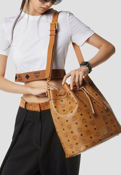 Femme portant un t-shirt court blanc et un pantalon noir, tenant un sac à main en cuir marron clair de la marque MCM avec motif logo et bandoulière réglable.