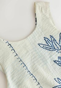 Tissu blanc sans manches avec des lignes de quadrillage bleu clair et un motif floral bleu brodé près de l'encolure et de la zone des épaules.
