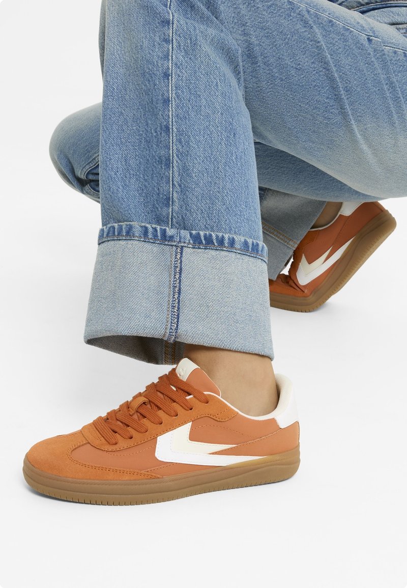 Baskets en daim orange et cuir avec accents blancs et semelle en caoutchouc gomme. Modèle à lacets avec matière texturée. Jean en denim visible.