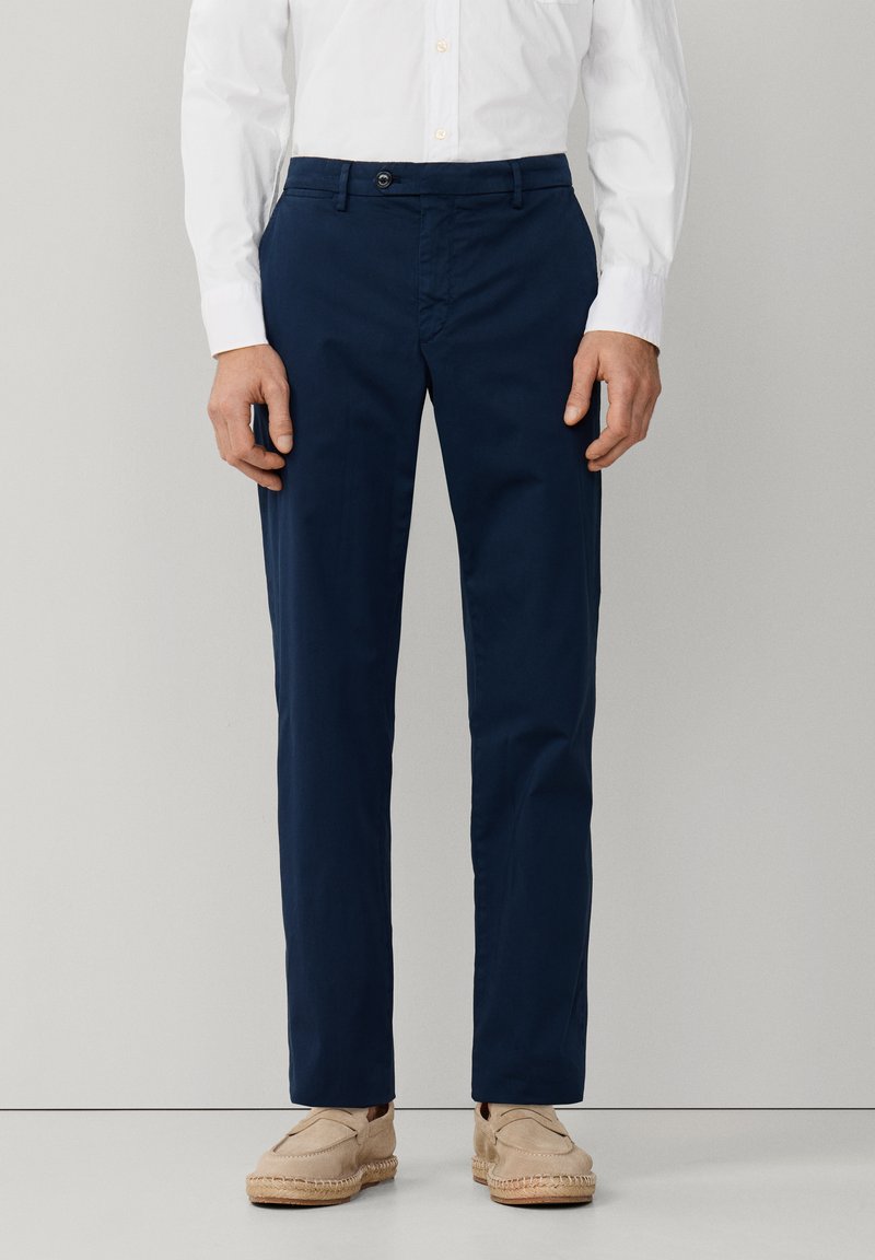 Homme portant un pantalon bleu marine ajusté, une chemise blanche boutonnée et des mocassins beiges décontractés, debout devant un fond gris uni.