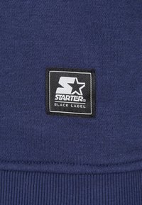 Parche del logotipo Starter Black Label en blanco y negro cosido sobre un tejido de punto de color azul marino texturizado.