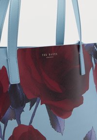 Zils sintētisks tote soma ar lielu rozes printu sarkanā un tumši zilā krāsā. Zelta "Ted Baker London" logotips centrā priekšpusē.