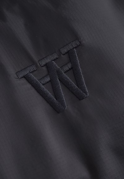 Tissu noir avec une surface lisse et légèrement texturée, ornée d'un logo brodé de la même teinte, représentant une lettre "W" stylisée.