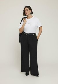 Camiseta blanca con texto impreso, pantalones de pierna ancha negros de cintura alta, cinturón negro con tachuelas de metal y zapatos negros de punta.