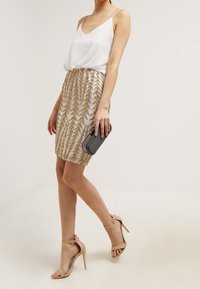 Haut en satin blanc sans manches, jupe dorée à sequins de longueur genou avec motif de feuilles, talons nus à lanières, et une pochette noire.