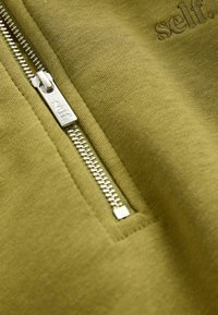 Tessuto verde oliva con una superficie liscia, dotato di una cerniera argento e del logo ricamato "self." vicino al bordo.