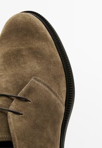 Scarpa in pelle scamosciata marrone con punta rotonda, suola in gomma nera e lacci neri sottili. Presenta cuciture visibili lungo il bordo e la tomaia.