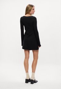 Vestido preto de mangas longas com silhueta ajustada, decote redondo e bainha rodada. Combinado com meias beige com bordas onduladas e sapatos pretos de salto bloco.