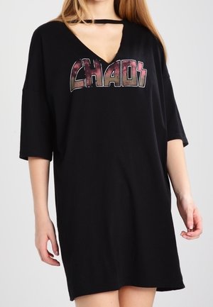 Femme portant une robe T-shirt noire oversize avec "CHAOS" imprimé sur le devant et un détail découpé au niveau de l'encolure.