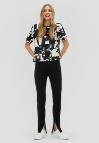 T-shirt à motif géométrique noir et blanc associé à un pantalon noir avec des fentes sur les côtés, porté avec des baskets blanches.
