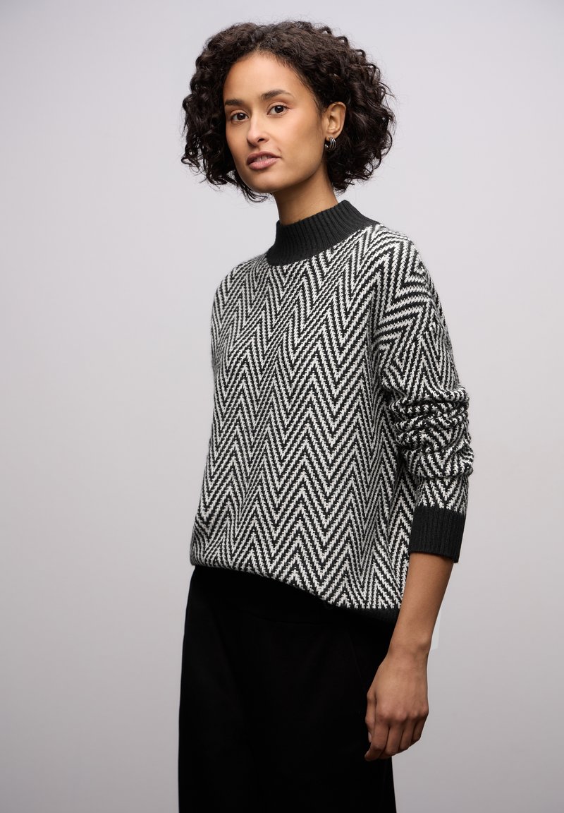 Femme aux cheveux bouclés et foncés portant un pull à motif zigzag noir et blanc et un pantalon noir, debout devant un fond uni clair.