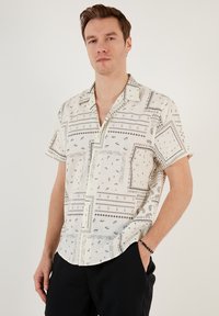 Camisa de manga corta abotonada en tela color crema con patrones negros, con un corte relajado, detalles en el cuello y secciones de patchwork distintas.