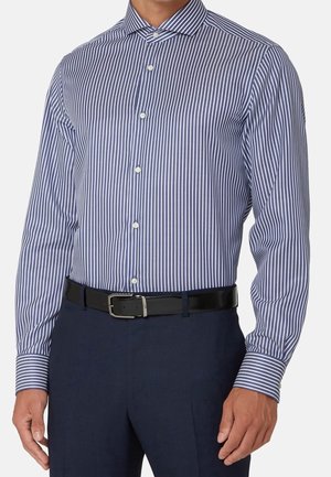 Homme portant une chemise à manches longues rayée verticalement bleu et blanc, rentrée dans un pantalon bleu foncé avec une ceinture noire.
