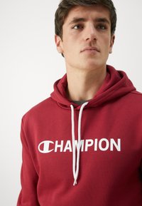 Rote Baumwollkapuze mit einem weißen "CHAMPION"-Logo. Verfügt über eine Kapuze mit Zugband und eine weiche Textur, die ein einfaches, lässiges Design präsentiert.