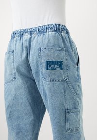 Pantalon en denim bleu clair avec taille élastique et plusieurs poches, une poche présentant un texte brodé bleu et un graphisme.
