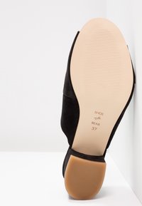 Chaussure en suède noire avec un bout rond et un talon bas. Semelle en caoutchouc lisse couleur tan avec texture nervurée. Nom de la marque et taille gravés sur la semelle.