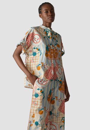 Woman wearing loose, colorful matching top and pants with abstract fruit and floral patterns on a light background, standing with hand in pocket.