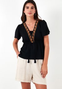 Zwarte blouse met korte pofmouwen, met oranje borduursel langs de V-halslijn en een trekkoorddetail met kwastjes in de taille.