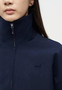 Femme portant une veste bleu marine zippée avec un col montant et un petit logo crocodile sur la poitrine gauche, sur un fond clair uni.