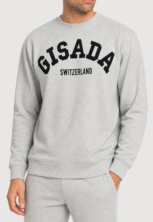 Homme portant un sweat-shirt gris clair et un pantalon assorti, le sweat-shirt porte la mention "GISADA" en grandes lettres noires et "SWITZERLAND" en dessous en plus petit.