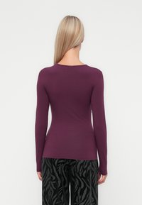Haut ajusté à manches longues en tissu violet profond, avec un col rond. Associé à un pantalon noir ample à motifs avec un design texturé.