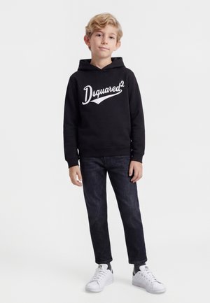 Lyshåret dreng iført en sort Dsquared2-hættetrøje, mørke jeans og hvide sneakers, stående foran en ensfarvet hvid baggrund.