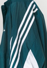 Veste bleu sarcelle foncé avec des rayures diagonales blanches sur la manche et un logo partiel, fabriquée en tissu léger.