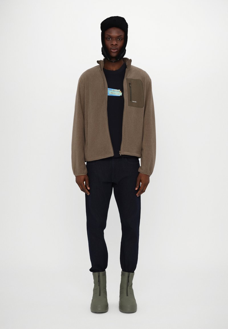 Braune Fleecejacke mit einer Fronttasche, über einem schwarzen T-Shirt getragen. Kombiniert mit dunklen Hosen und grünen Reißverschluss-Stiefeln.
