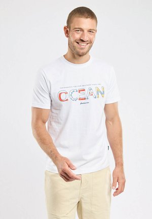 Lächelnder Mann, der ein weißes T-Shirt mit buntem "OCEAN"-Schriftzug und beige Hosen trägt, steht vor einem schlichten weißen Hintergrund.