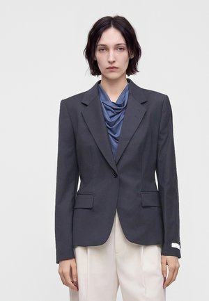 Frau mit kurzen dunklen Haaren, die einen dunkelgrauen Blazer über einem blauen, drapierten Oberteil und hellbeigen Hosen trägt, stehend vor schlichtem Hintergrund.