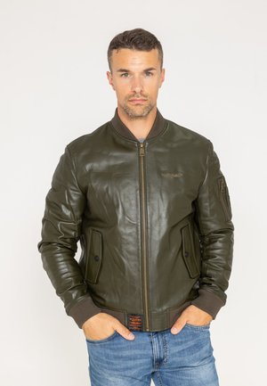Mężczyzna z krótkimi włosami, ubrany w oliwkowozieloną skórzaną kurtkę bomberkę i niebieskie dżinsy, stojący z rękami w kieszeniach na białym tle.