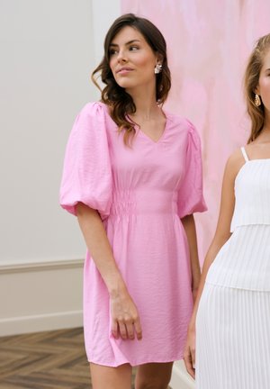 Jeune femme aux cheveux ondulés portant une robe rose à manches bouffantes, debout à côté d'une autre en robe plissée blanche à l'intérieur.